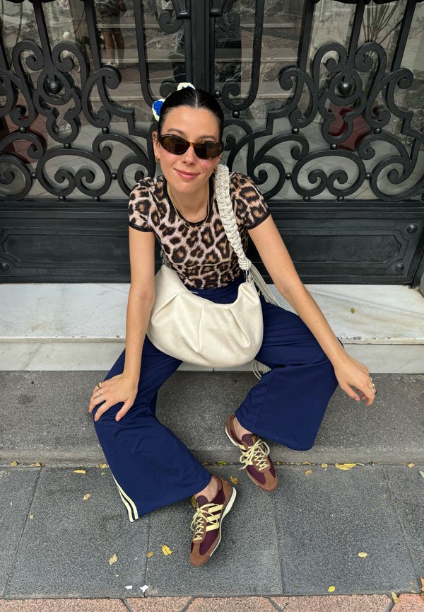 Topp med leopardmønster, marineblå vide bukser, beige slouchy veske og brune sneakers med gule snørebånd, sittende mot en dekorativ svart port.