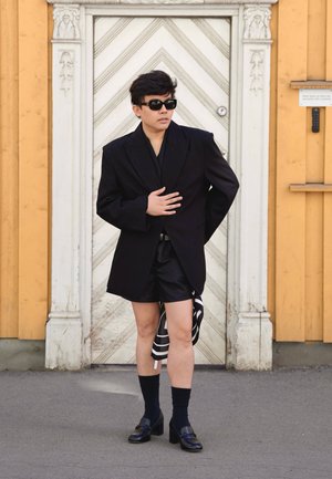 Persona con una blazer negra oversize, pantalones cortos negros, gafas de sol, calcetines negros y zapatos de tacón, de pie frente a una puerta blanca ornamentada.