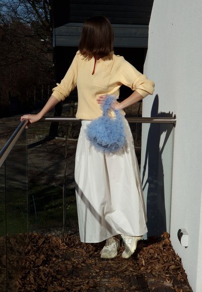 Suéter amarillo acanalado, falda larga blanca y bolso de volantes azul claro. Zapatillas blancas completan el conjunto. Al aire libre con follaje debajo.