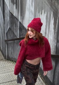 Pull rouge duveteux court à manches longues avec un bonnet assorti, associé à un pantalon camouflage, sur un fond urbain texturé.
