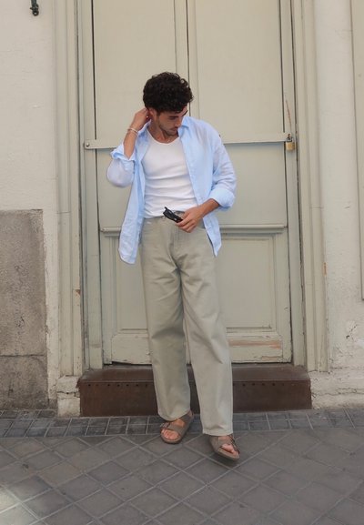 Camisa de botón azul claro sobre una camiseta blanca, combinada con pantalones caqui claros y sandalias marrones, de pie frente a una puerta de madera cerrada.