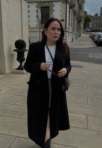 Czarny długi płaszcz z guzikami, biała koszula pod spodem, beżowa spódnica, czarna torebka, trzymając okulary przeciwsłoneczne, stojąc na wybrukowanej ulicy.