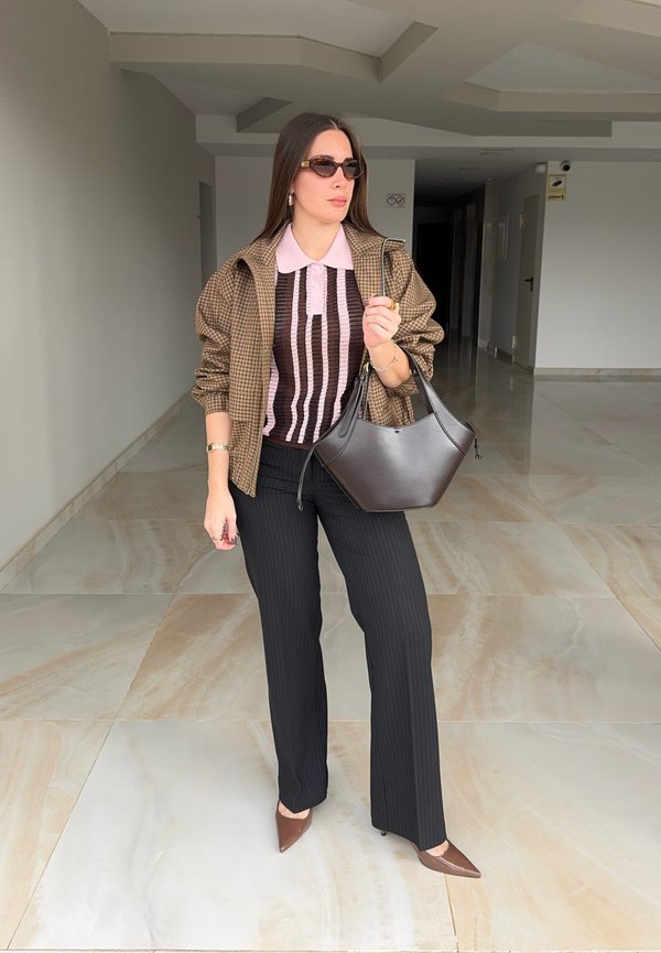 Kobieta w okularach przeciwsłonecznych, w brązowej kurtce w kratę, pasiastym różowo-czarnym topie, czarnych spodniach, brązowych szpilkach, niosąca ciemnobrązową torebkę w pomieszczeniu.