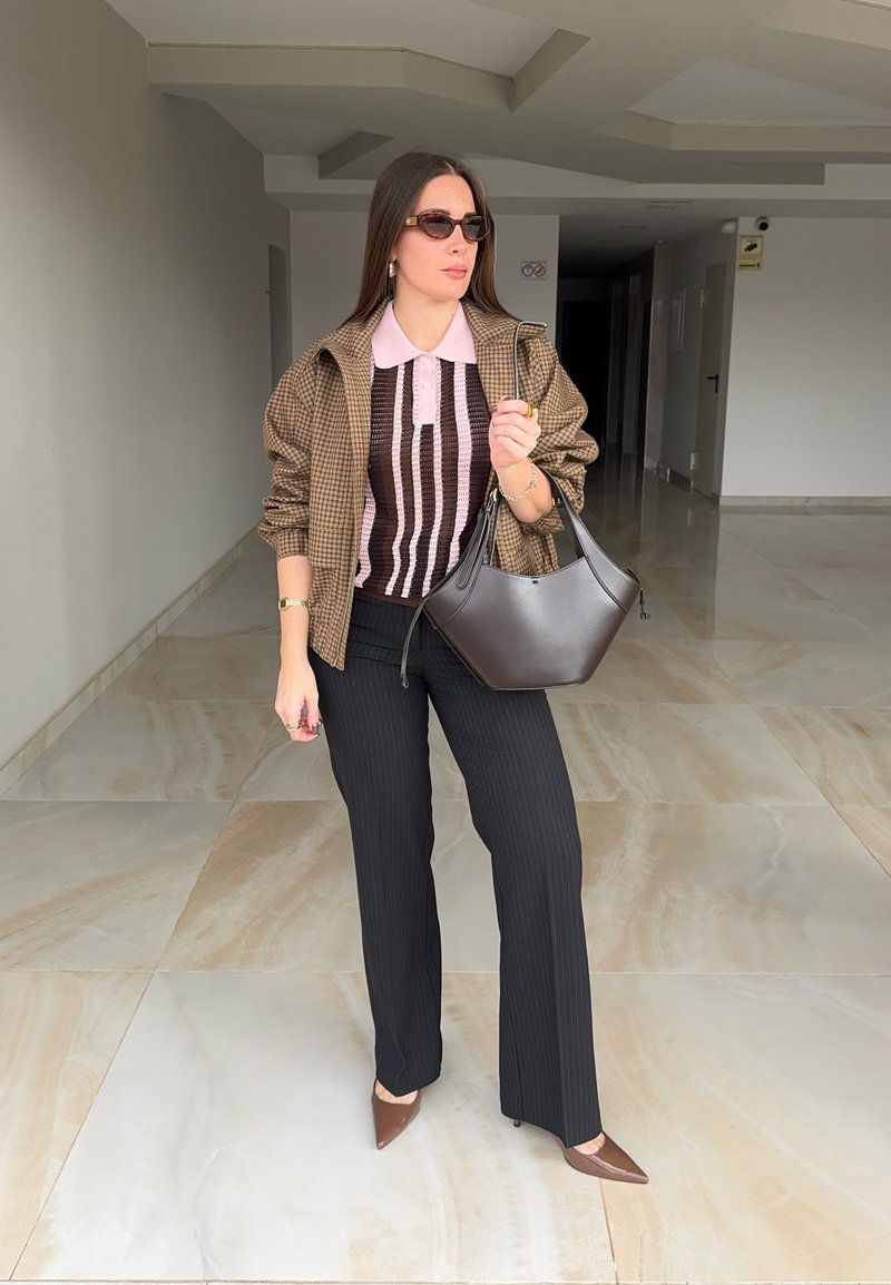 Femme portant des lunettes de soleil, une veste à carreaux marron, un haut rayé rose et noir, un pantalon noir, des talons marron, portant un sac à main marron foncé à l'intérieur.