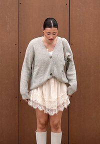 Jasnoszara, dziergana kardiganka z guzikami z przodu, zestawiona z kremową sukienką wykończoną koronką. Białe botki do kolan dopełniają stylizację.