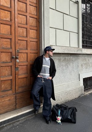 Hombre con gafas de sol y gorra apoyado en una puerta de madera, vestido con abrigo oscuro y vaqueros, con bolso negro y llavero de peluche en la acera.