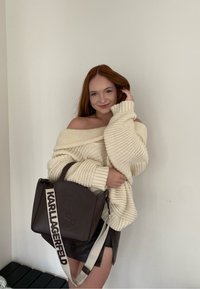 Suéter de punto crema con diseño de hombros al aire, combinado con un bolso marrón oscuro con logo y correa ajustable. Falda de cuero visible.