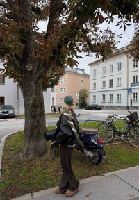 Una persona con i capelli lunghi indossa una giacca di pelle nera e un berretto verde, in piedi vicino a un albero e a biciclette parcheggiate, con uno scooter nelle vicinanze.