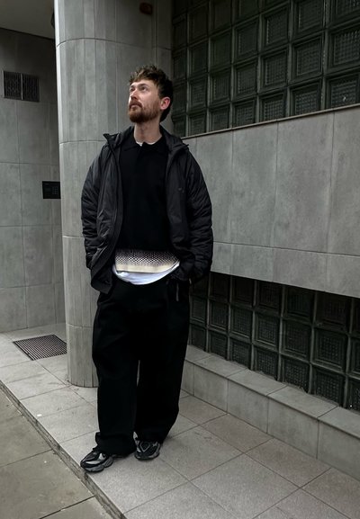 Chaqueta acolchada negra, camisa negra y pantalones negros holgados; camiseta blanca con un patrón texturizado; zapatillas deportivas grises; de pie contra una pared de azulejos.