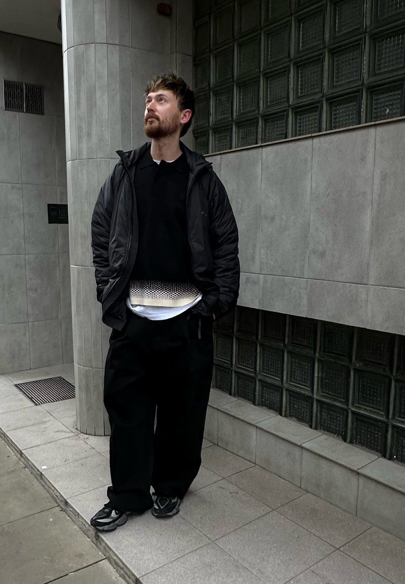 Schwarze gesteppte Jacke, schwarzes Hemd und übergroße schwarze Hose; weißes Unterhemd mit strukturiertem Muster; graue Turnschuhe; steht vor einer gekachelten Wand.