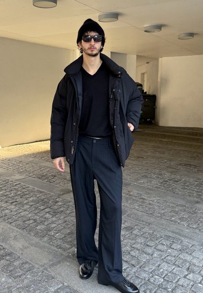 Chaqueta acolchada negra sobre una camiseta negra con cuello en V, combinada con pantalones de rayas finas y zapatos negros. Los accesorios incluyen gafas de sol y un gorro.