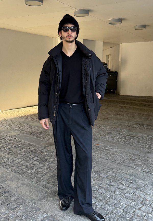 Casaco acolchoado preto sobre uma camisa preta com decote em V, combinado com calças riscas de giz e sapatos pretos. Acessórios incluem óculos de sol e um gorro.