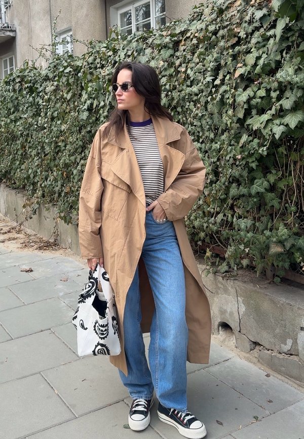 Kvinne iført beige trenchcoat, stripete skjorte, blå jeans, svarte joggesko og solbriller, holder en svart-hvitt mønstret veske, står på fortauet.
