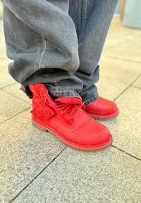 Sarkanas zamšādas potītes zābaki ar apaļu purngalu, gumijas zoli un auklām. Tajos ir pievienota "50" logotipa etiķete. Kopā ar brīviem pelēkiem džinsa biksēm.