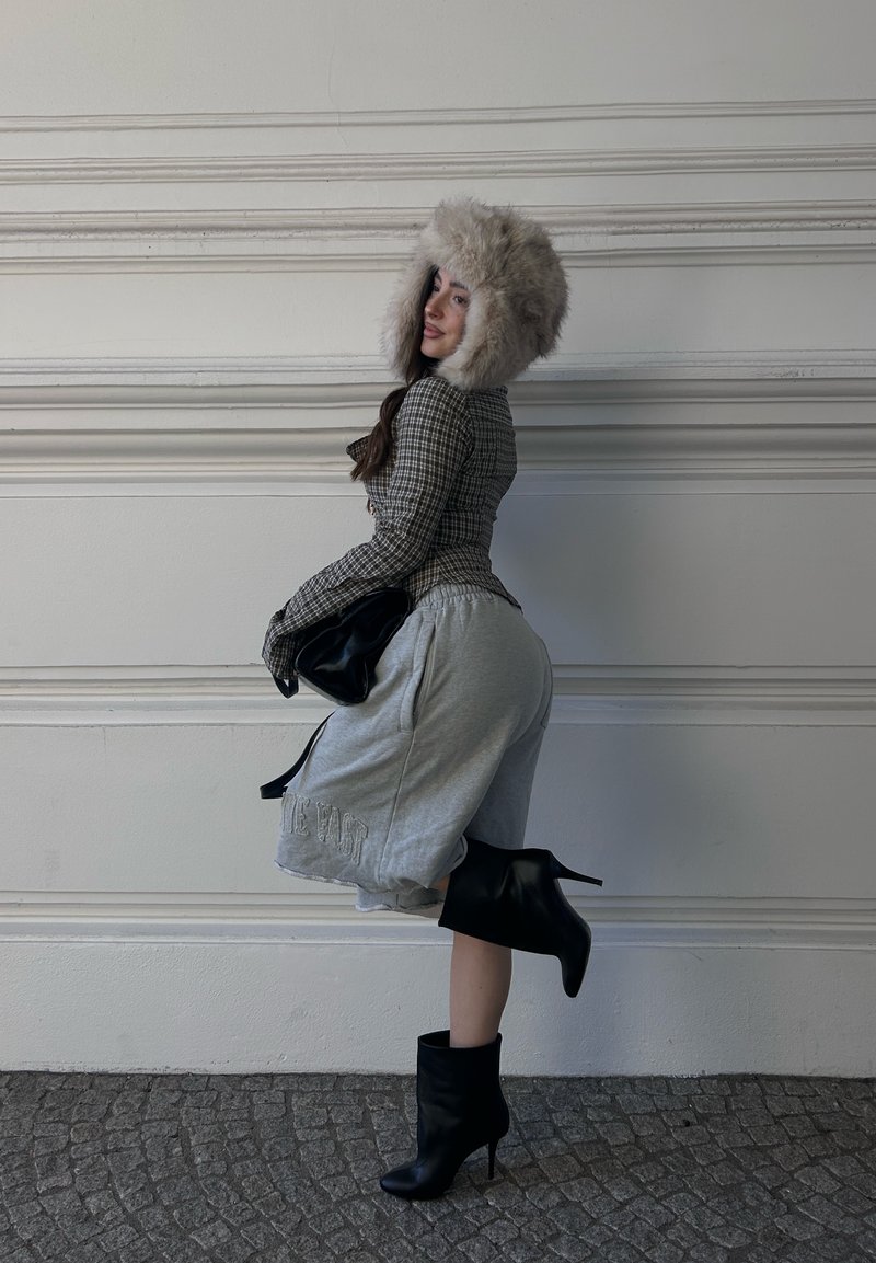 Femme portant un chapeau en fourrure, une veste à carreaux, un short gris jusqu'aux genoux et des bottes noires à talons hauts, posant contre un mur blanc.