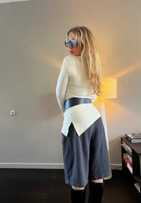 Femme aux longs cheveux blonds portant de grandes lunettes de soleil, un haut crème avec une ceinture noire, un short ample foncé et des bottes noires, debout à l'intérieur.