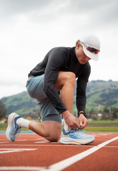 Atleta atándose las zapatillas de running HOKA de color azul claro con acentos naranjas en una pista roja, vistiendo una camiseta de manga larga negra y pantalones cortos grises.