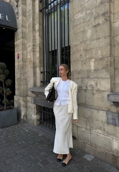 Blazer blanco sobre una blusa blanca de punto trenzado y falda crema. Bolso negro y zapatos planos de color claro con puntas oscuras. Fondo de pared de piedra.