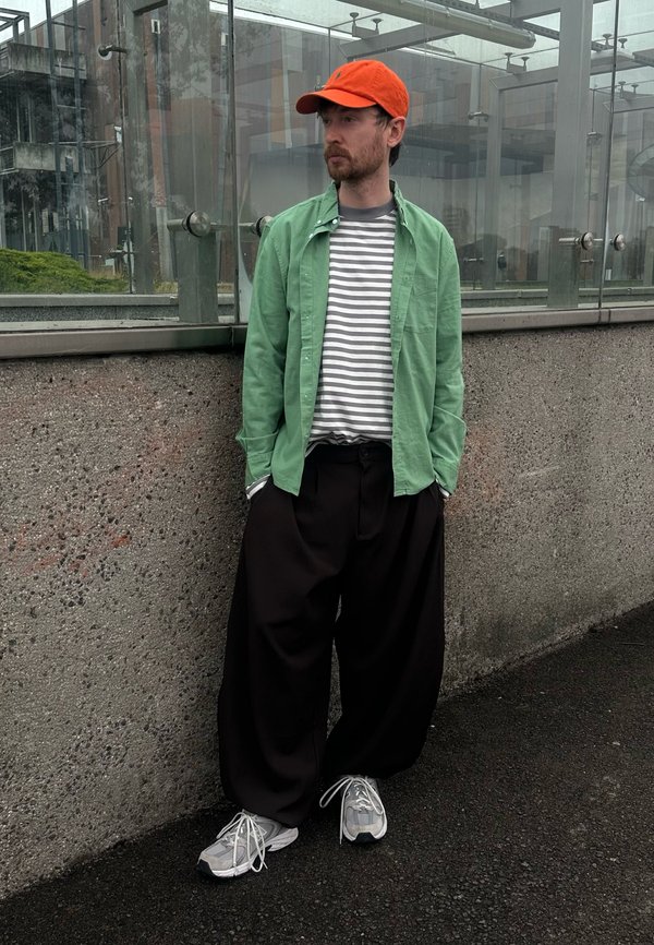Hombre con gorra naranja, camisa verde, camiseta de rayas, pantalones negros anchos y zapatillas grises, de pie junto a una pared de hormigón cerca de paneles de vidrio.