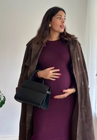 Crna torba sa struktuiranim dizajnom, koju nosi osoba u bordo haljini s rebrastim uzorkom i smeđem baloneru, držeći se za trbuh.