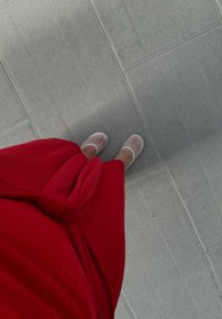 Robe rouge, à mi-mollet, en maille côtelée avec un détail noué à la taille. Portée avec des sandales plates doux et translucides. Sol texturé gris en dessous.