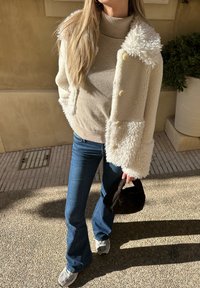 Casaco de fleece em creme com colarinho texturizado e três botões sobre um suéter de gola alta bege. Jeans azul de boca de sino e mala preta completam o conjunto.