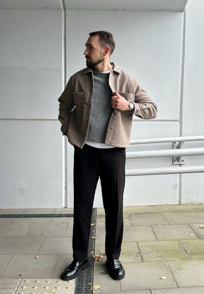 Chaqueta marrón de botón sobre un suéter gris, combinada con pantalones negros y mocasines negros, de pie sobre una superficie de baldosas con un fondo de pared gris.