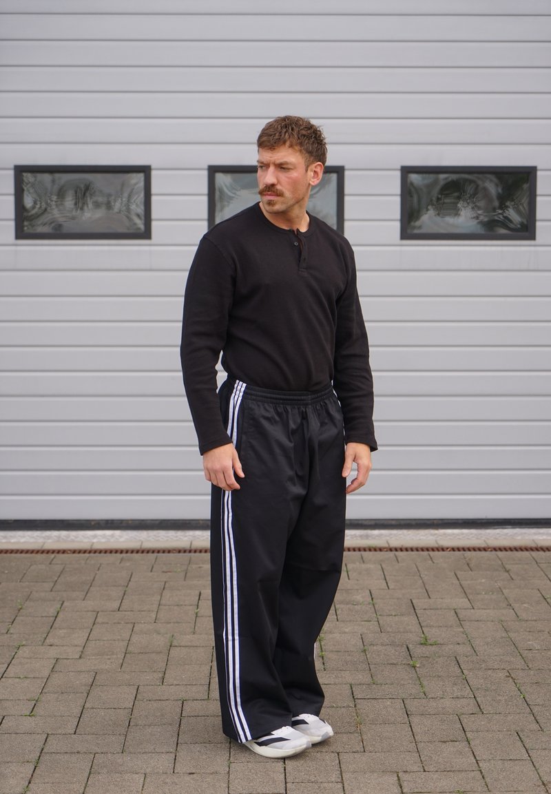Homme avec moustache portant une chemise noire à manches longues et un pantalon noir à rayures blanches, debout sur un sol pavé devant un mur gris.