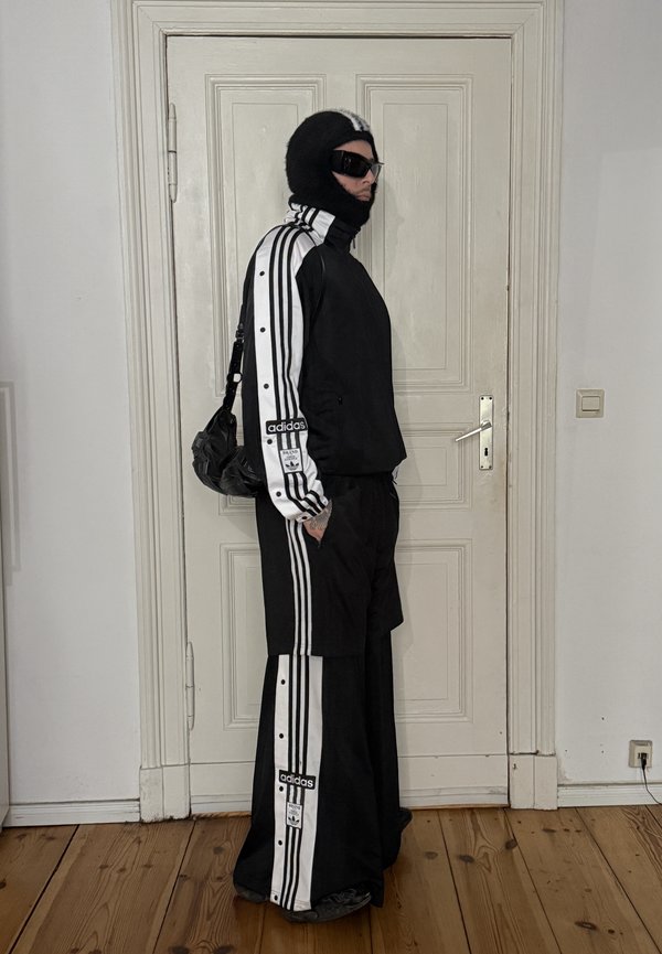 Човек в страничен изглед, облечен в черно-бял екип на Adidas с райета, черна балаклава, слънчеви очила и носещ черна чанта в стая.