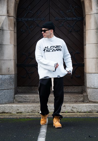 Suéter blanco oversized con el texto "LOVE SCARS", detalles desgastados, pantalones de chándal negros con franjas laterales y botas amarillas. Gorro negro y gafas de sol.