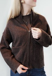 Hnedý zapínací sveter na zips z mäkkej textúrovanej tkaniny, s dvoma prednými vreckami a rebrovanými manžetami, nosený cez tmavý top.