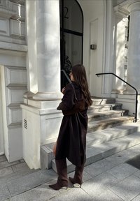 Hnědý oversize trenčkot, hnědé sukní boty sahající ke kolenům se špičatými špičkami a tmavá ramenní taška. Hladká textura a strukturovaný design.