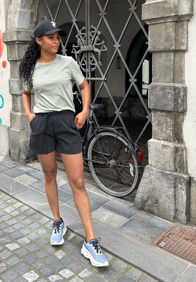 Pantalones cortos negros y una camiseta verde claro con el texto "Peak Performance". Zapatillas gris-azul con una suela blanca gruesa. Gorra negra. Fondo de pared de piedra.