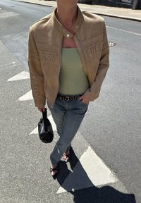 Jaqueta moto em camurça tan com texto em relevo, blusa verde em malha, jeans azul claro, mala preta e sandálias pretas com tiras.