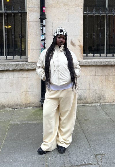 Chaqueta puff de color crema con un cuello alto, pantalones anchos de color crema y un sombrero de patrón en blanco y negro. Cabello trenzado suelto.