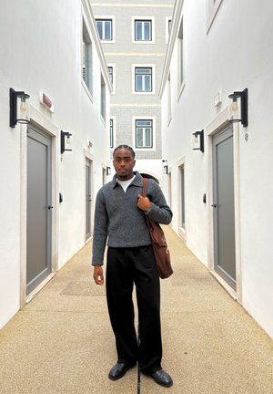 Hombre con el pelo trenzado, vestido con un suéter gris y pantalones negros, de pie en un pasillo estrecho entre edificios blancos con múltiples puertas.