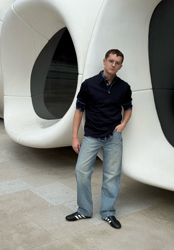 Man met bril, gekleed in een zwarte trui, opgerolde geruite blouse, losse spijkerbroek en sneakers, leunt tegen een grote witte moderne architectonische structuur.