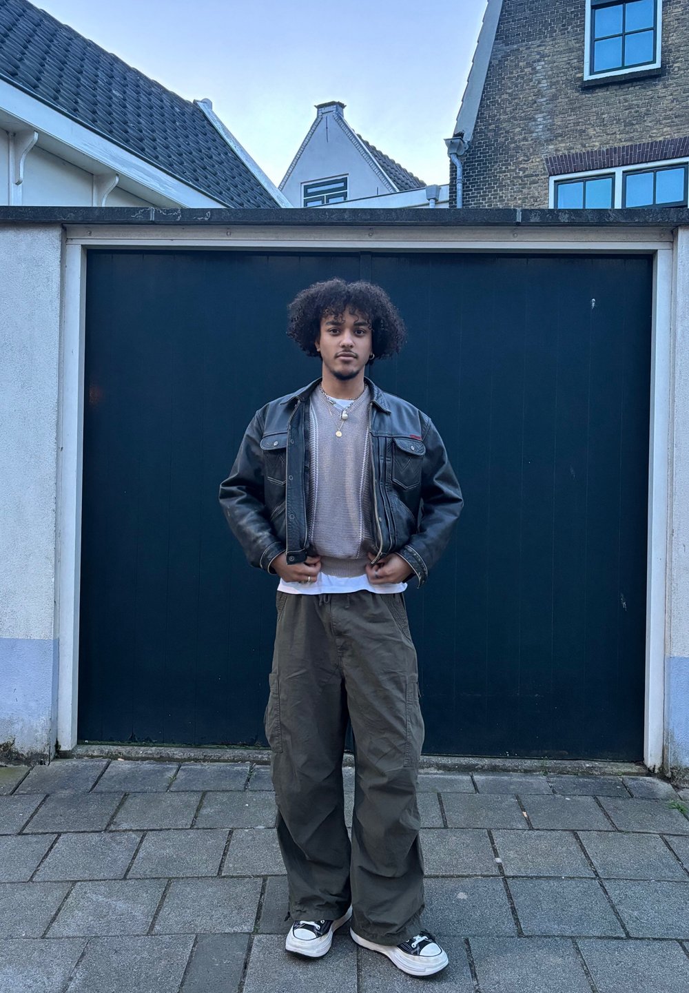 Fiatal férfi, göndör hajjal, fekete bőrkabátban, bézs pulóverben, cargo nadrágban és sportcipőben áll egy térkövezett úton, egy sötét garázs előtt.