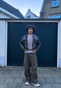 Fiatal férfi, göndör hajjal, fekete bőrkabátban, bézs pulóverben, cargo nadrágban és sportcipőben áll egy térkövezett úton, egy sötét garázs előtt.