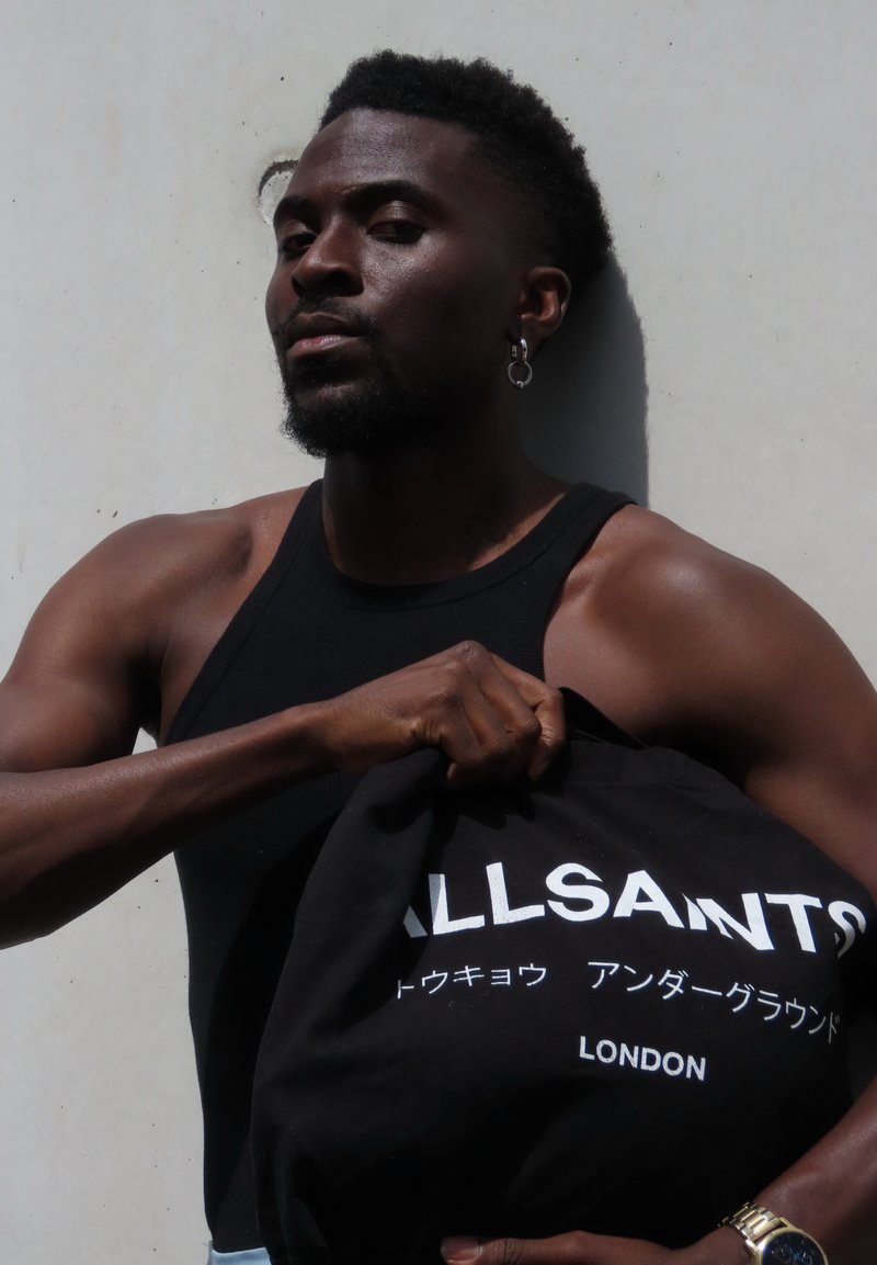 Homme musclé en débardeur noir tient un sac tote noir ALLSAINTS contre un mur clair, portant une boucle d'oreille et une montre.
