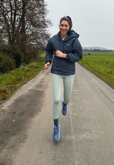 Mujer con chaqueta oscura y leggings claros corriendo por un camino rural pavimentado, rodeada de campos de hierba y árboles sin hojas bajo un cielo gris.