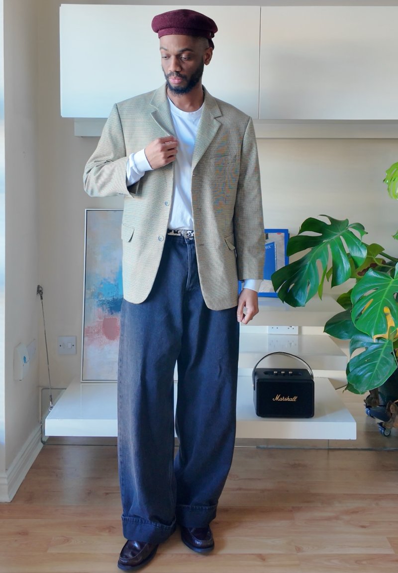 Checkered beige blazer, white shirt, dark wide-leg jeans, and maroon beret. Black speaker and green plant in background.