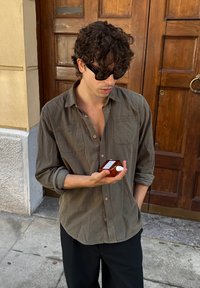 Bruine overhemd met opgerolde mouwen, houdt een heldere glazen fles met een witte dop vast. De achtergrond toont houten deuren.