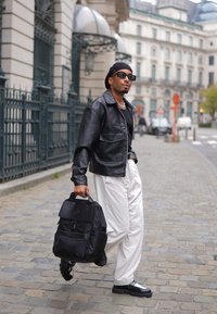 Jachetă din piele neagră cu un design cu guler, pantaloni albi cu pantaloni largi și un rucsac negru cu multiple buzunare, purtat casual pe o stradă.