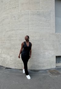 Svart tettsittende topp med dyp V-hals og lang, stram skjørt, paret med hvite joggesko. Bakgrunnen har en betongvegg og et vindu.