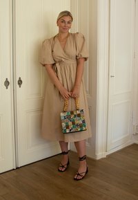 Mujer en un vestido midi de mangas acampanadas color beige, sosteniendo un bolso a cuadros con asas gruesas, vistiendo sandalias negras de tiras y joyería dorada, de pie en el interior.