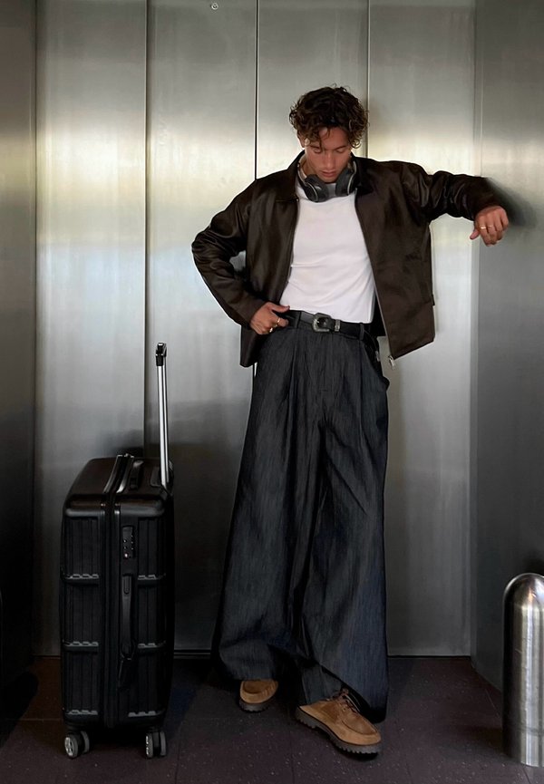 Negro maleta de rodillo a la izquierda; persona con una chaqueta de cuero marrón, camisa blanca, pantalones oscuros anchos y zapatos marrones, en un ascensor.
