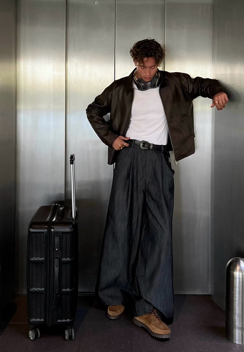 Valise à roulettes noire à gauche ; personne portant une veste en cuir marron, une chemise blanche, un pantalon large noir et des chaussures marron, dans un ascenseur.