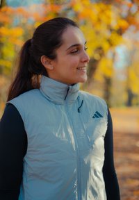 Chaleco sin mangas azul claro con cuello alto, cremallera frontal y detalles en negro. El chaleco presenta un logo de Adidas en el lado izquierdo.