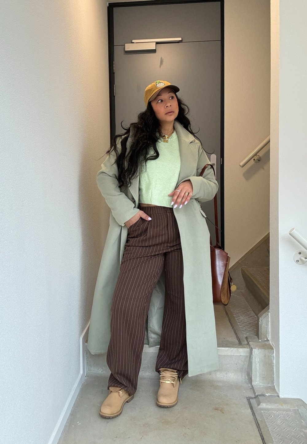 Lange lichtgroene jas over een muntgroene trui, bruine gestreepte broek en tan laarzen. Accessoires omvatten een tan pet en een bruine tas.